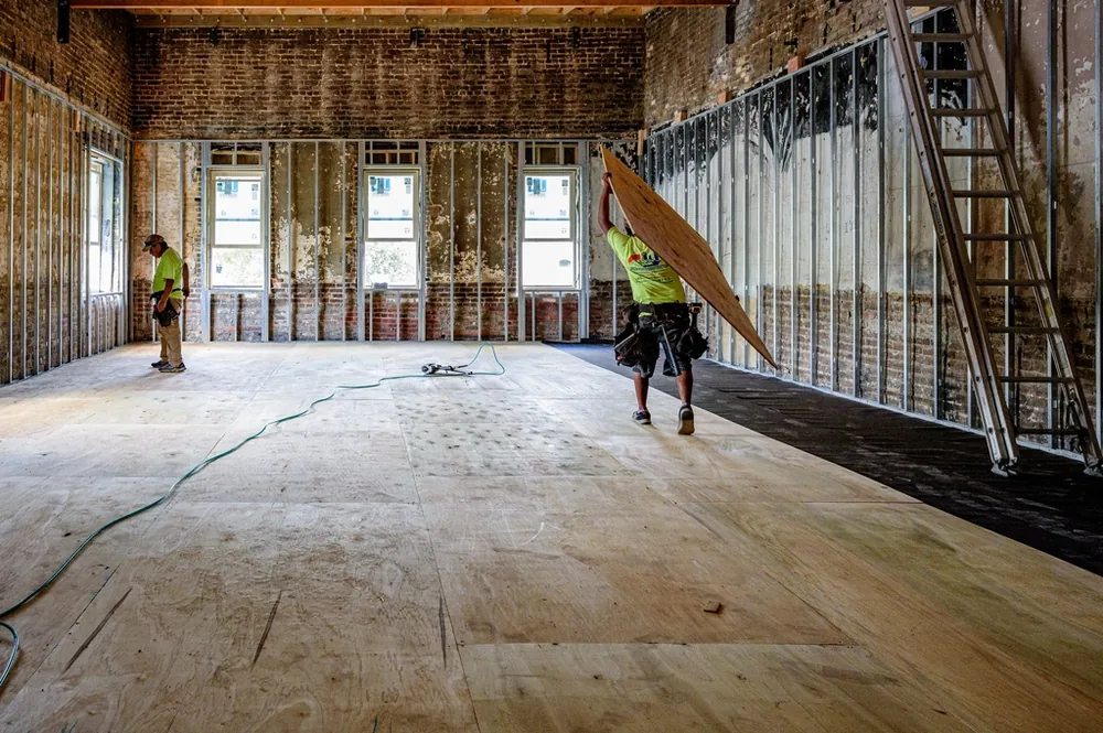Worker carrying a sheet of drywall across a gutted room